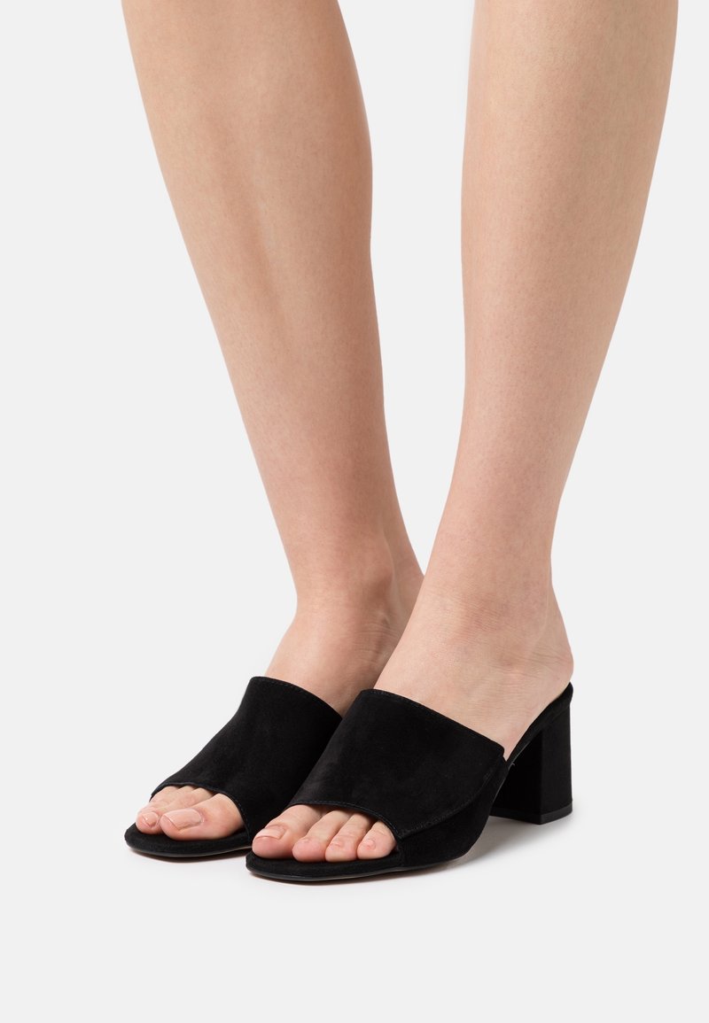 Pieds portant des sandales à talons blocs ouverts en suède noir sur un fond clair uni.