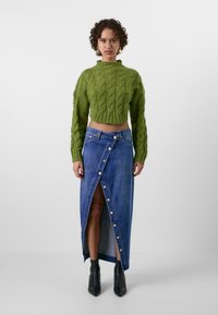 Pull cropped en tricot vert associé à une jupe maxi en denim bleu avec une fente avant et des détails de boutons dorés. Bottes noires à chevilles.