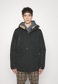 Black jacket with a hood, featuring large front pockets, zippered chest pocket, and tan collar lining. Worn over a gray hoodie.