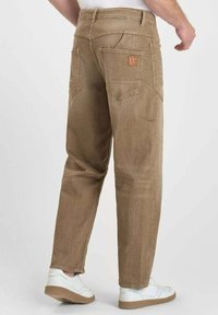 Homme portant un pantalon marron ample avec plusieurs poches arrière et des baskets blanches, se tenant devant un arrière-plan clair.