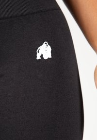 Črne športne hlače s teksturirano tkanino in majhnim belim logotipom dveh stiliziranih figur na spodnji strani.