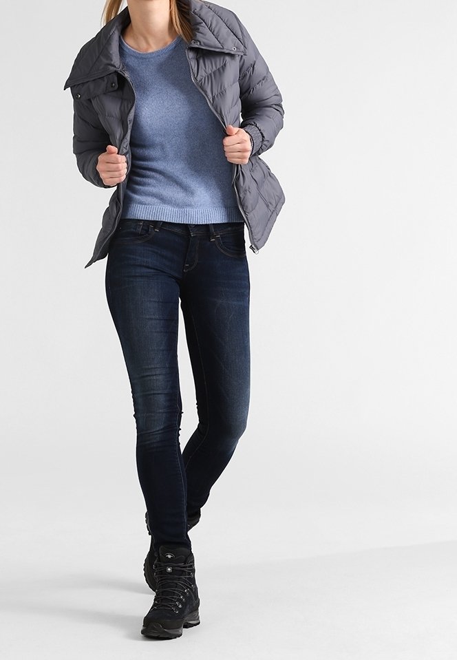 Femme portant une doudoune grise par-dessus un pull bleu, un jean skinny foncé et des bottes de randonnée noires, marchant sur un fond blanc uni.