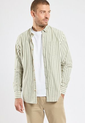 Jeune homme portant une chemise à boutons beige et blanche à rayures verticales par-dessus un t-shirt blanc avec un pantalon beige, regardant sur le côté.