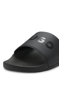 Μαύρη σαγιονάρα με υφασμένο πέλμα και φαρδιά λουρί που φέρει το έντονο λογότυπο "HUGO" σε γυαλιστερά μαύρα γράμματα.