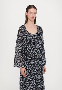 Abito nero floreale con maniche lunghe e trasparenti e scollatura ampia, cingetto in vita con una cintura nera caratterizzata da accessori metallici.