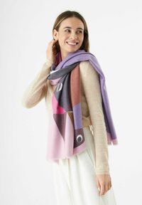 Écharpe à motif géométrique coloré dans des teintes de violet, rose et gris. Texture douce avec des bords frangés. Portée en drapé sur un pull léger.
