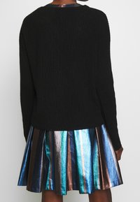 Černý žebrovaný svetr s dlouhými rukávy v kombinaci s metalickou skládací sukní s vertikálními pruhy v modré, zelené a bronzové barvě.