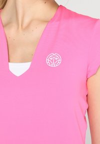 Kortärmad, v-ringad, klar rosa skjorta gjord av lent tyg med en vit logotyp på bröstet. Inre lager synligt vid halsringningen.