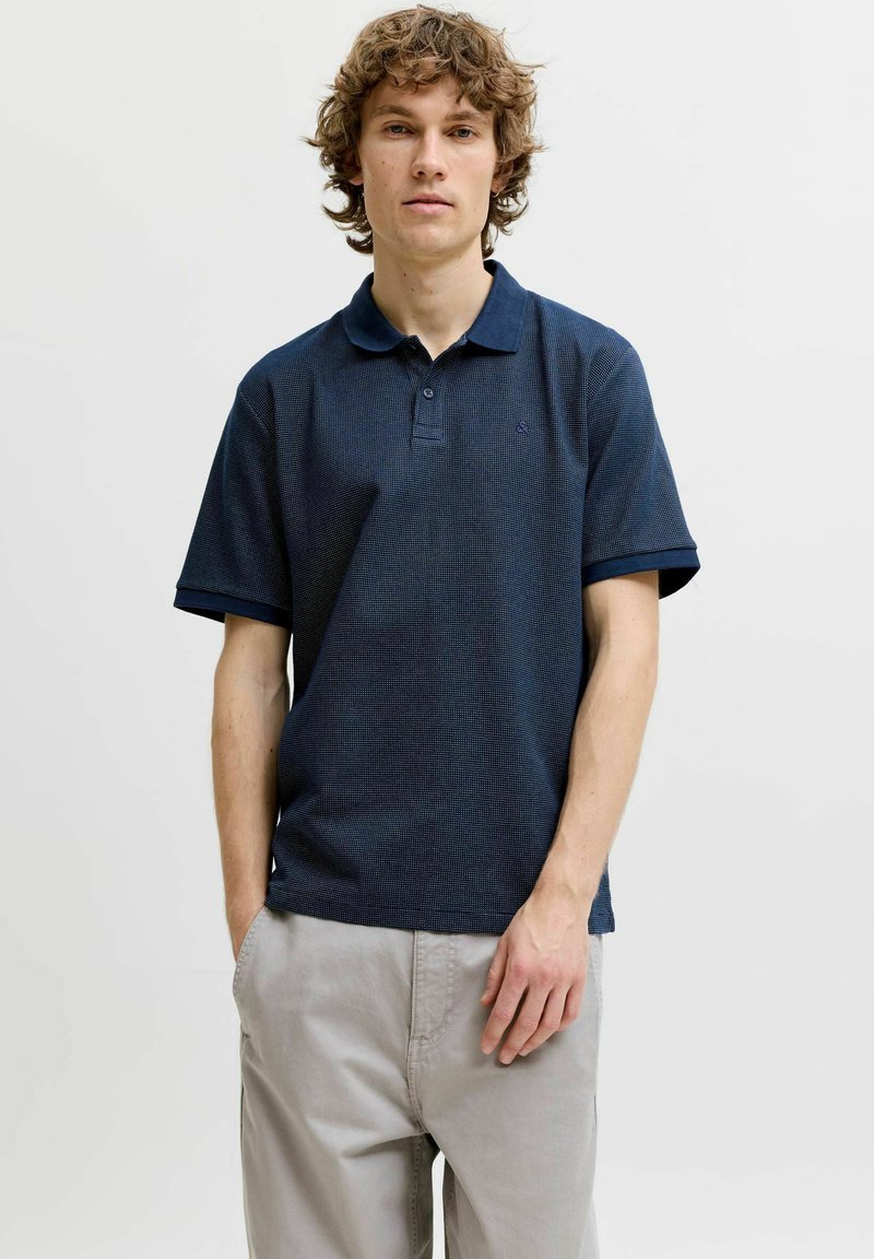 Jeune homme aux cheveux bouclés portant un polo bleu marine à manches courtes et un pantalon beige clair, debout sur un fond blanc uni.