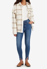 Camisa de franela a cuadros en crema con líneas azules y naranjas, camiseta blanca, jeans skinny oscuros con aberturas laterales y mocasines marrones.