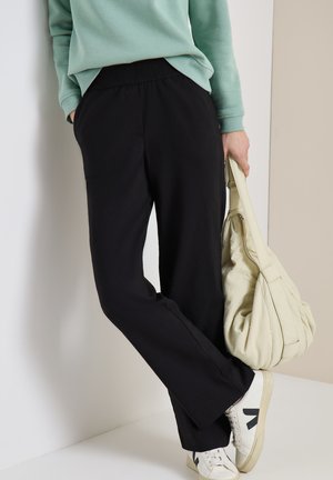 Persona con pantalones negros de cintura elástica, zapatillas blancas con logo negro, sudadera verde menta, sosteniendo bolso beige holgado.