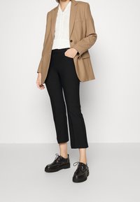 Blazer marron, haut en tricot blanc, pantalon court noir et chaussures à lacets noires. Tissu lisse, coupe ajustée, détails subtils comme des poches et des lacets.