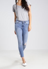 Camiseta gris de manga corta combinada con unos jeans ajustados de color azul claro que presentan una rodilla desgastada, terminando con unos planos blancos. Diseño simple y casual.