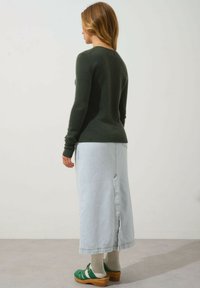 Donkergroene trui met lange mouwen, gecombineerd met een lichtblauwe denimrok met een uitsparing aan de achterkant. Groene klompen en witte geribde sokken maken de look af.