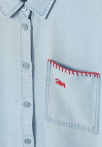 Chemise bleu clair à boutons avec des détails de couture rouges et une petite broderie rouge de homard sur la poche poitrine.