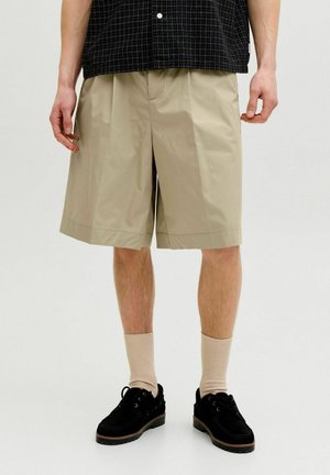 Homme portant un short plissé beige, des chaussures noires, des chaussettes beiges et une chemise à carreaux noire, debout sur un fond blanc uni.