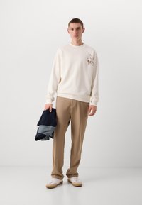 Krem sweatshirt med tegneserie-design på brystet, beige bukser og hvite sneakers med beige detaljer. Holder en sammenfoldet mørk denim.