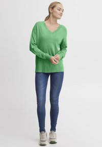Pulover verde cu maneci lungi, de tip V, cu o croială lejeră, asociat cu blugi albaștri mulati și adidași deschiși la culoare. Textură netedă cu manșete cu nervuri.