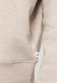 Acercamiento a la manga de un suéter de punto beige con costura visible y una pequeña etiqueta de tela blanca en la costura lateral.