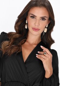 Schwarzes Wickelkleid mit langen Ärmeln, goldene strukturierte Ohrringe und ein großer goldener Ring. Haare weich gewellt gestylt. Neutraler Hintergrund.