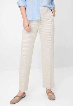 Woman wearing beige patterned straight-leg trousers, light blue shirt with rolled sleeves, and beige flat shoes.