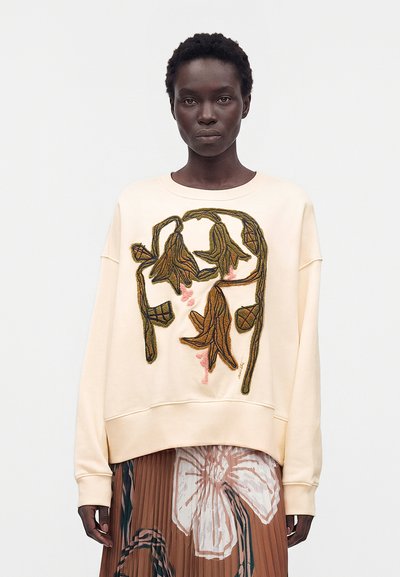 Femme aux cheveux courts et bouclés portant un sweat crème avec une large broderie florale marron et verte et une jupe plissée avec un imprimé fleuri surdimensionné.