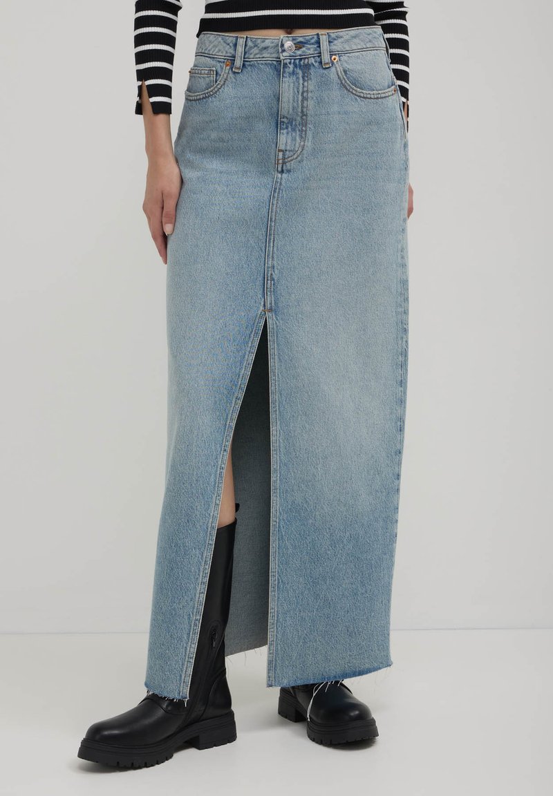 Jupe en denim bleu clair avec une fente à l'avant, coupe droite et ourlet effiloché, assortie à des bottes noires jusqu'aux genoux.