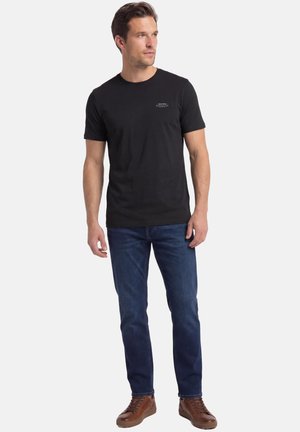 Man staat in een zwart T-shirt met korte mouwen, blauwe spijkerbroek en bruine leren sneakers, en kijkt naar links.