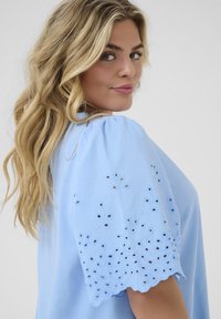 Lichtblauwe katoenen blouse met puffmouwen en geborduurde bloemenpatronen. Heeft uitsnijdingen langs de mouwranden. Soepele textuur.