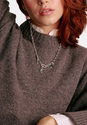 Silberne Halskette mit Anhängern: eine Fledermaus, ein Schwert und ein florales Muster. Das Model trägt einen grauen, strukturierten Pullover über einem weißen Hemd mit Kragen.