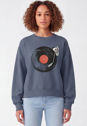 Frau mit lockigem Haar, die einen blauen Sweatshirt mit einer Grafik einer schwarzen Vinyl-Schallplatte und eines Plattenspielers auf der Vorderseite trägt.
