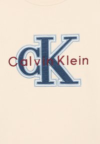 Tela de color beige claro con un gran logo bordado multicolor: "CK" en azul marino y "Calvin Klein" en color burdeos. Textura suave.