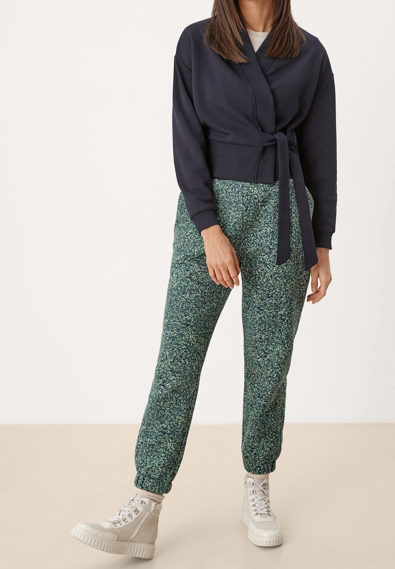 Femme portant un pull cache-cœur bleu marine, un pantalon de jogging vert à motifs et des baskets montantes beige clair, debout devant un fond uni.