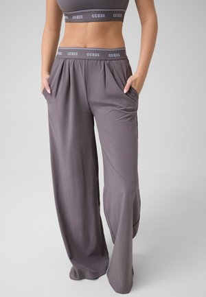 Frau trägt lockere graue Hose und passendes bauchfreies Oberteil, beide mit elastischem „GUESS“-Bund, Hände in den Taschen, vor schlichten Hintergrund.