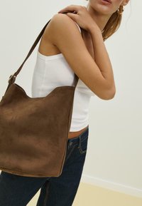 Borsa a spalla in camoscio marrone con un design morbido e sformato e un bordo superiore curvo. La tracolla è liscia e resistente.