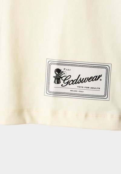 Cream-colored cotton t-shirt featuring a black graphic label with "Godsweat" and a character illustration. Smooth texture, short sleeves.