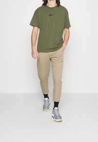 Olivově zelené bavlněné tričko s malým černým logem, kombinované s khaki zúženými kalhotami s logem na kapse a šedými teniskami se žlutými akcenty.