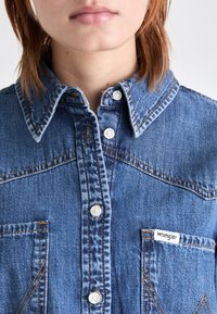 Keskmise sinise tooniga denimipäevik, valgete nööpidega, terava kraega, rinna taskuga ja kontrastsete õmbluse detailidega. Brändi silt: Wrangler.