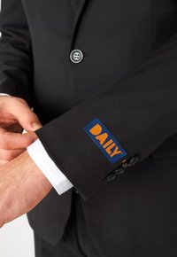 Chaqueta de traje negra con un parche de tela azul etiquetado "DAILY" en naranja, con un puño de camisa blanca visible en el borde de la manga.
