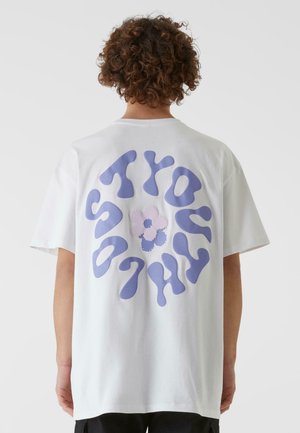 Hvit bomulls t-skjorte med et stort lila smeltende tekstdesign på ryggen, med en liten blomstergrafikk i midten.