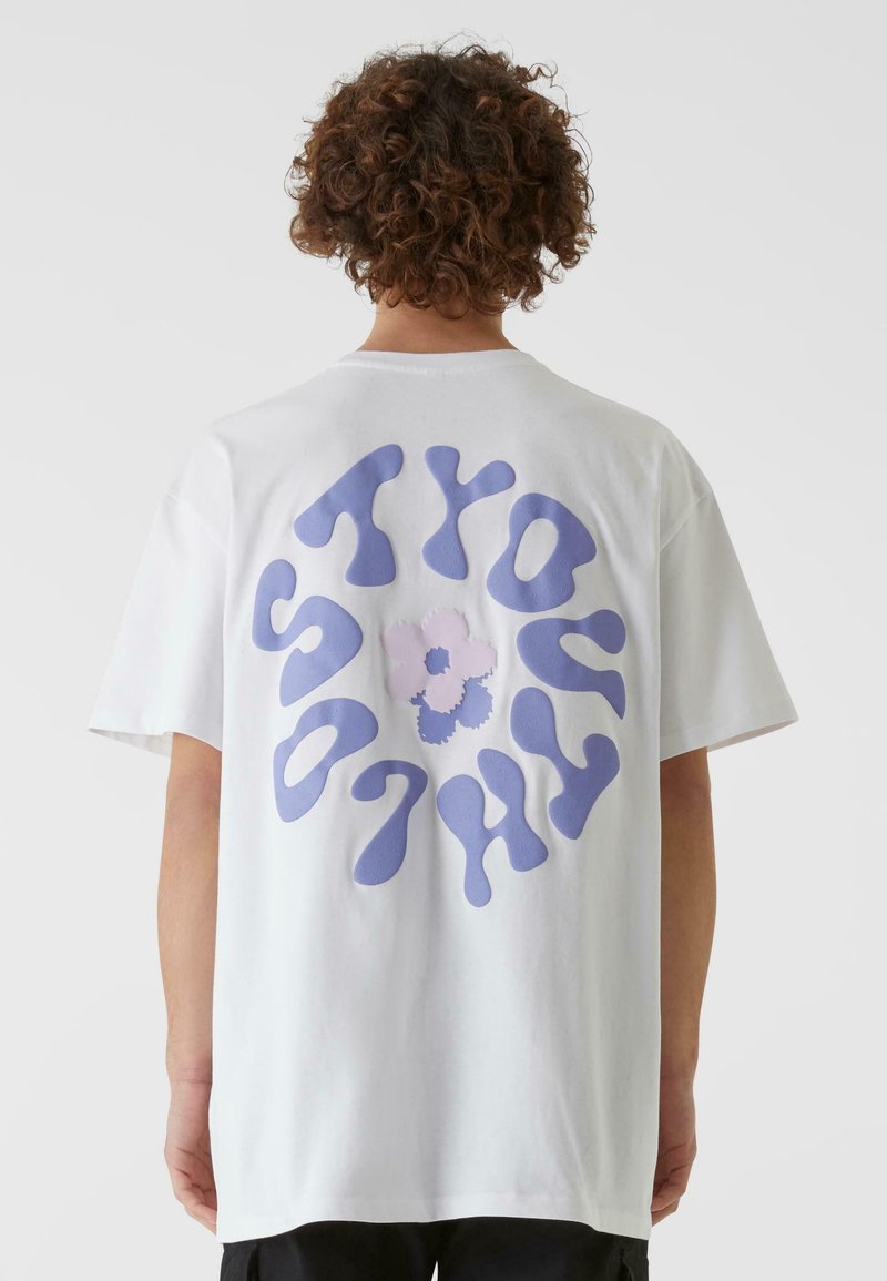 Hvit bomulls t-skjorte med et stort lila smeltende tekstdesign på ryggen, med en liten blomstergrafikk i midten.