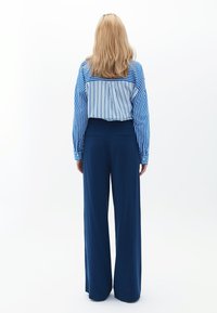 Blauw-wit gestreept overhemd met knopen en lange mouwen, gecombineerd met wijde marineblauwe broeken. Zachte textuur, ontspannen pasvorm en minimale details.