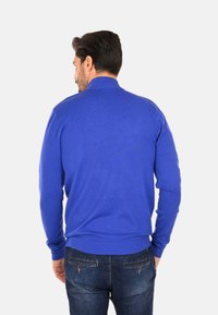 Maglione blu lavorato a maglia con colletto alto, polsini e orlo a coste. Indossato con jeans in denim scuro, presenta un design semplice e testurizzato.