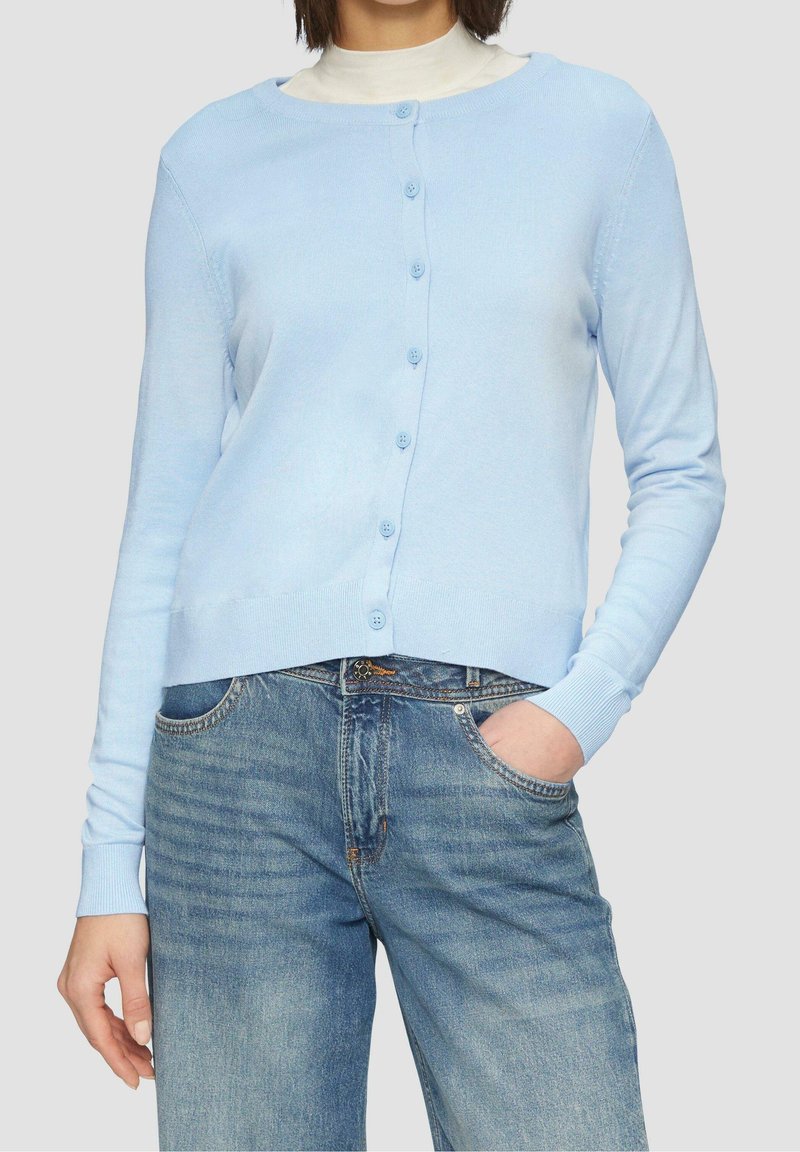 Femme portant un cardigan bleu clair boutonné sur un haut blanc associé à un jean en denim bleu, avec une main dans la poche.