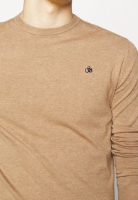 Beige pletený svetr s kulatým výstřihem, na levé straně s malým tmavě modrým logem "&". Rukávy jsou mírně nařasené na manžetách.