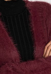 Burgundy fluffig cardigan med en texturerad, lång svart ribbad polotröja under. Cardiganen har en mjuk, fluffig yta.