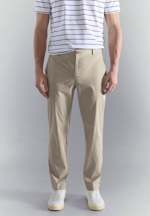 Uomo con pantaloni beige a taglio dritto, scarpe da ginnastica bianche e camicia bianca a maniche corte con sottili strisce orizzontali nere.