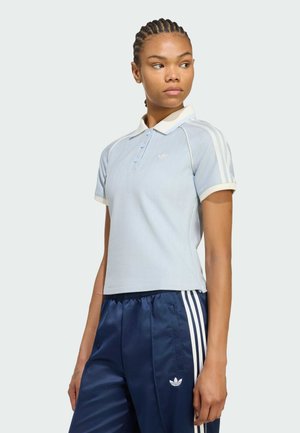 Žena s copy, která má na sobě světle modré polo tričko s krátkým rukávem a tmavě modré sportovní kalhoty s bílými pruhy, dívající se stranou.