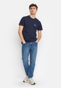 Marineblauw katoenen t-shirt met korte mouwen en een kleine print aan de voorkant, gecombineerd met blauwe spijkerbroek en lichtgekleurde sneakers.