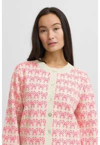 Cardigan en tricot avec un fond crème présentant un motif de nœuds roses répété, un col rond et une fermeture à boutons à l'avant avec des boutons clairs.